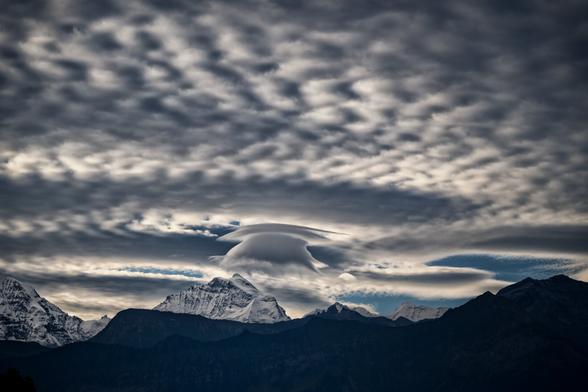Dramatische Berglandschaft mit schneebedeckten Gipfeln im Hintergrund. Besonders auffällig sind die außergewöhnlichen Wolkenformationen am Himmel - glatte, linsenförmige Wolken, die wie geschichtete Scheiben oder Wellen aussehen. Die Berge erscheinen in bläulichen Tönen durch die atmosphärische Perspektive, während der bewölkte Himmel in verschiedenen Grau- und Weißtönen gehalten ist.