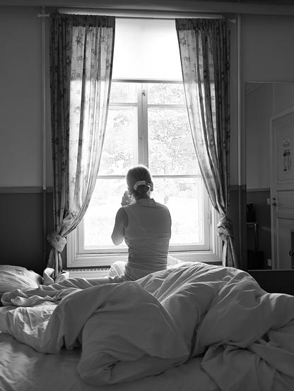 Black and white photo of a woman sitting on a bed facing a window and doing her makeup. White duvet is in the foreground in an untidy pile. The woman wears white tank top and her hair is in a bun. Her back is towards the photographer and she is sitting so that the curtains are framing her. On the right side of the wall there is a mirror, that shows a door.