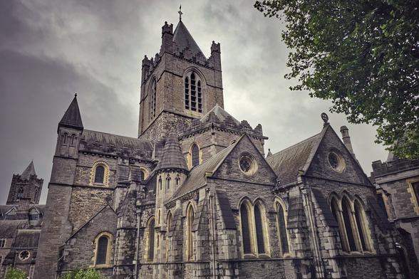 A low-angle shot of Christ Church Cathedral in Dublin, Ireland, under a dramatic, cloudy sky. The historic stone building with its tall, dark grey steeple and intricate gothic arches fills the frame, with a branch of a leafy tree visible in the top right corner.