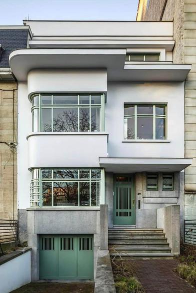 art deco house in Brussels, streetview.
Garage with green doors on the left (lower etage).
Stairs on the right going to green deco door.
Windows and rooftops rounded shaped and with horizontal elements.
The house is white. Accent are green.