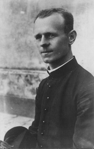 Black and white photo of a seated priest wearing a clerical collar and buttoned-up attire.