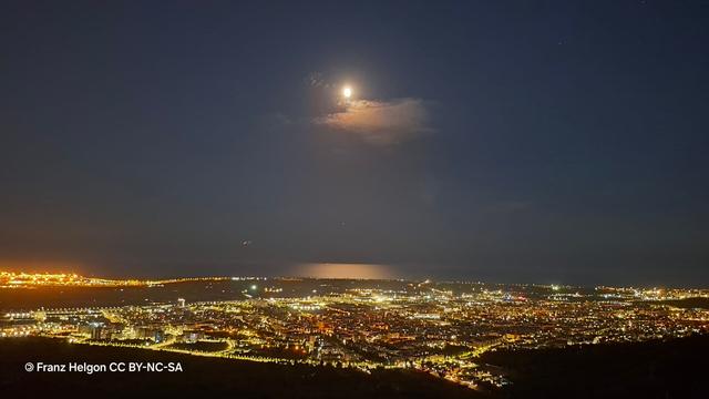 Imatge nocturna. Es veu una ciutat plena de llums sota un cel seré i la lluna plena al centre alt de la composició. Aquesta lluna es reflecteix una mica en el mar.