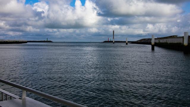 Color photo of a calm harbor entrance with green and red beacons marking the way to the open sea under a partly cloudy sky. Gentle ripples cover the water’s surface, while concrete piers and breakwaters frame the scene.