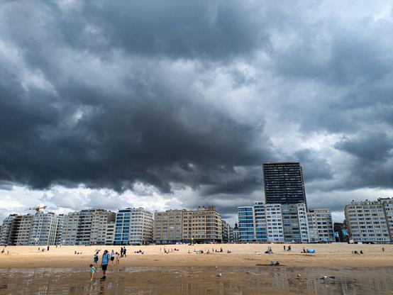 Color photo of a beach scene with people walking and relaxing on the sand under a dramatic sky filled with dark, heavy clouds. In the background, a row of modern apartment buildings and a tall, slightly leaning skyscraper dominate the skyline.