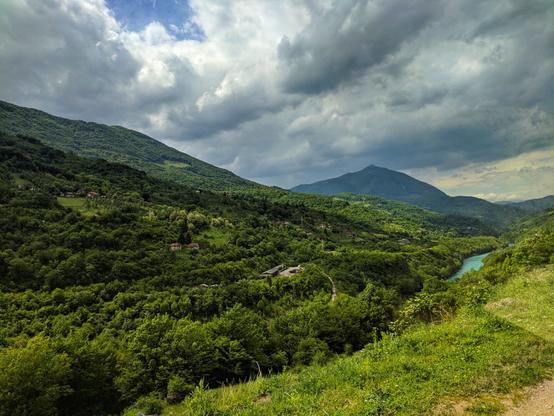 A panoramic shot of the Drina River valley in Bosnia and Herzegovina, taken from a high vantage point. The turquoise river winds its way through a lush green landscape of forested hills and mountains. In the foreground, the vibrant green grass and trees lead the eye down into the valley. Small houses and a road are dotted across the hillsides. The sky above is dramatic, with a mix of bright blue patches and large, heavy white and grey clouds, suggesting a recent or impending change in the weather. The overall impression is one of peaceful, remote natural beauty.