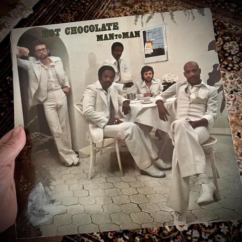 The image features the cover of a music album titled "Man to Man" by Hot Chocolate. It shows five men dressed in white suits, seated and standing in a stylish interior setting, with a table set for tea. The atmosphere is elegant, all the members looking at the camera.