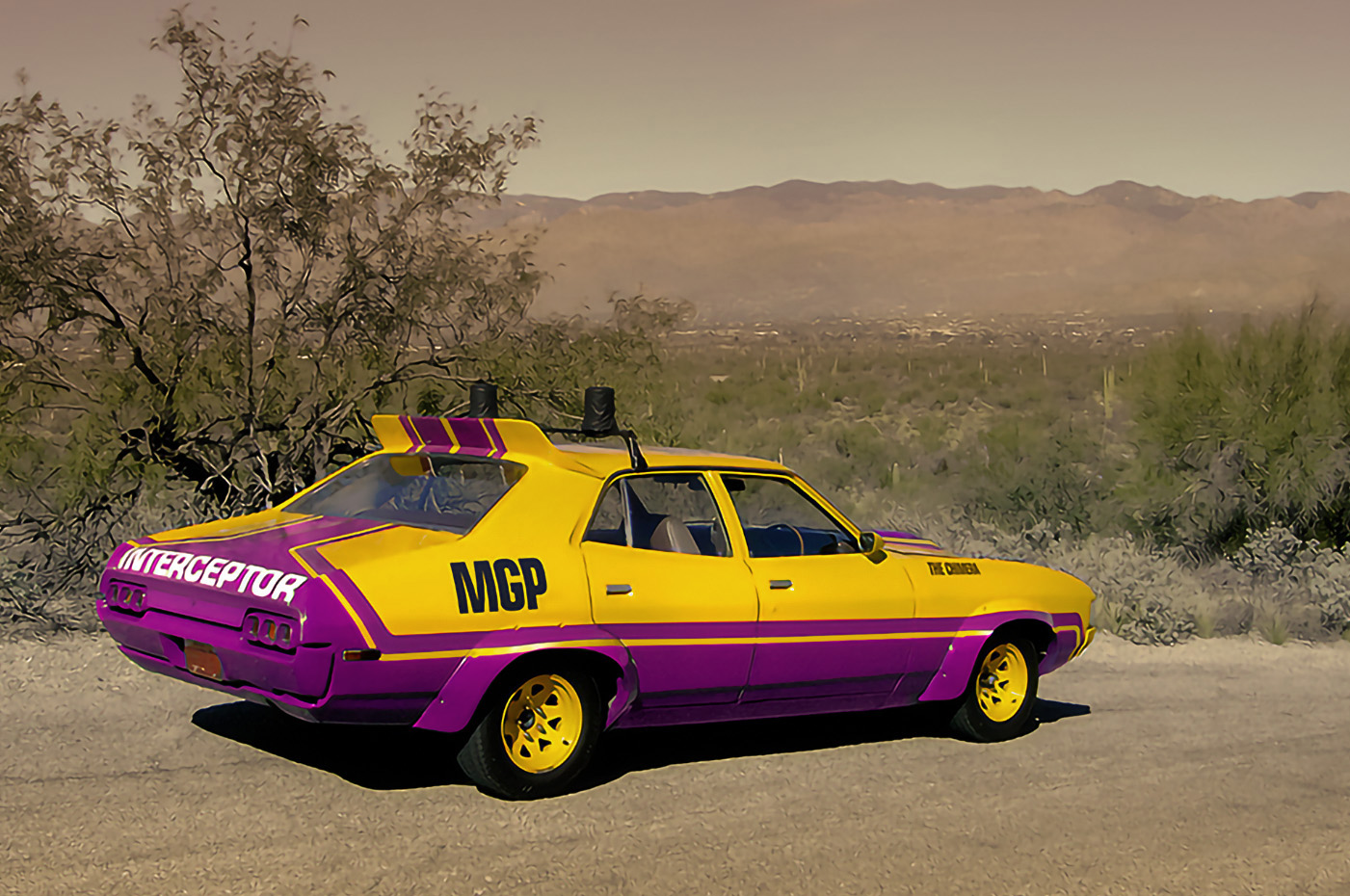 A photo of a mad max MFP interceptor in Yellow and Purple colours, those of the intersex flag. It's a 1972 XA Falcon sedan parked on a gravel road. In the background are hills and a dull brownish sky.