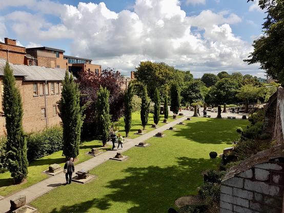 This image captures a serene, well-maintained garden flanked by tall, slender cypress trees lining a stone pathway. The lush green lawn is dotted with small stone artifacts, possibly historical remnants, adding an ancient charm to the scene. A few people stroll leisurely along the path, enjoying the sunny weather under a sky filled with fluffy white clouds. On the left, old brick buildings with traditional windows contrast with a more modern structure in the background. To the right, a stone wall partially covered with greenery adds texture and depth, while the garden opens up to a more expansive area with circular stone arrangements and benches where people are seated, likely enjoying the peaceful ambiance. In the distance, dense trees create a natural boundary, blending shades of green that complement the vibrant lawn. The play of light and shadow across the scene enhances the tranquil, picturesque atmosphere of this beautiful outdoor space.