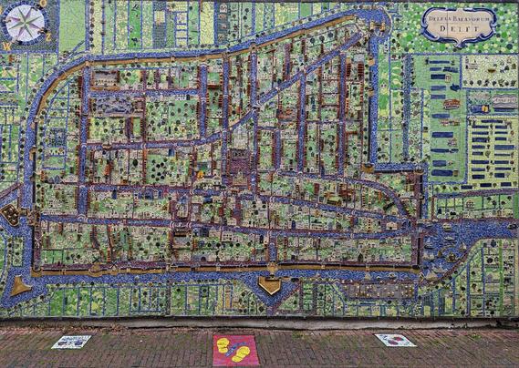 A vibrant, large-scale mosaic map of the city of Delft, Netherlands, is mounted on a brick wall. The map is highly detailed and three-dimensional, depicting the city's historic canals (shown in blue), streets, and buildings in colourful ceramic tiles and other materials. The surrounding landscape, including fields and waterways, is also rendered in mosaic. A compass rose is visible in the top-left corner, and a cartouche in the top-right corner reads "DELFTIA BATAVORUM DELFT". Below the mosaic, on the red brick pavement, are three smaller square tiles, each with a different design.