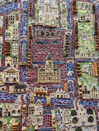 A close-up shot of a section of a large, three-dimensional mosaic map of Delft. The image focuses on the city's central area, showcasing highly detailed, relief-style ceramic tile representations of famous buildings. The 'Stadthuys' (City Hall) and 'Nieuwe Kerck' (New Church) are prominently featured, crafted in white and gold-toned tiles. Surrounding them are smaller, individual buildings, some with written street names such as 'MARCKT VELT', 'VOLDERS GRACHT', and 'DRUDE'. The streets are depicted with red tiles, while the canals are rendered in blue and white tiles. The background is a mix of green and other colourful tiles, giving a sense of the surrounding landscape.