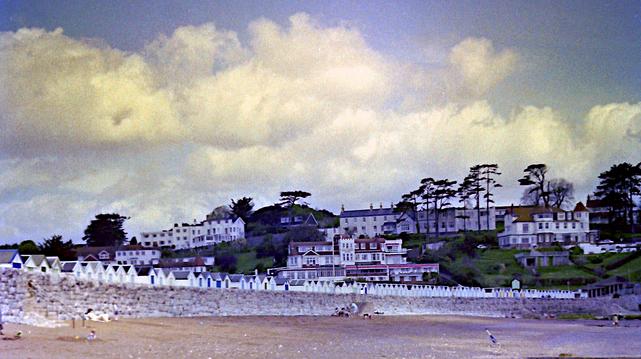 A traditional seaside resort, with a row of beach huts & hotels on a hill, behind.