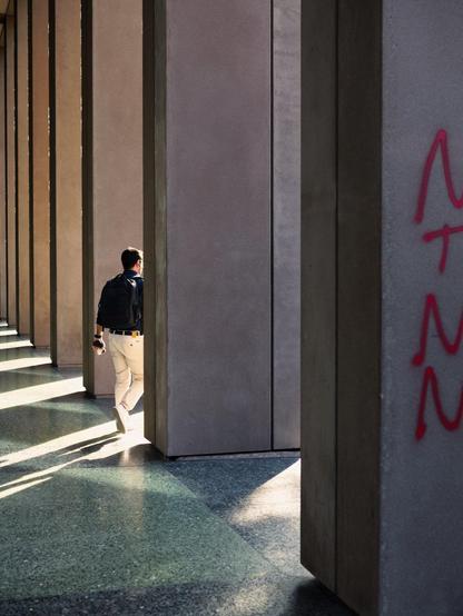 A young man walks away from us through tall, square columns. Sharp sunlight shines through the columns, creating a stark contrast in the scene.