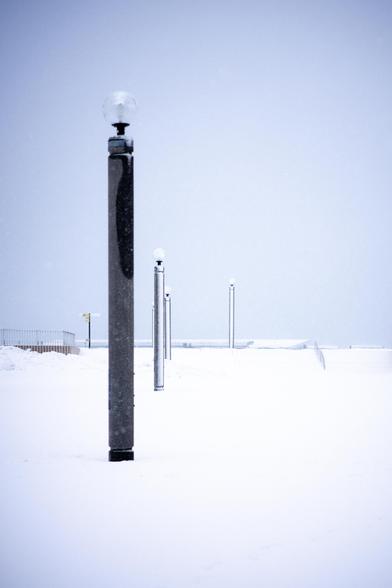 Four lamp posts line a snow-covered pathway under an overcast sky. The foreground features a close-up of the first lamp post, with the others receding into the background. Snow blankets the ground and enhances the monochromatic scene. A fence and distant structures are faintly visible on the left side.
