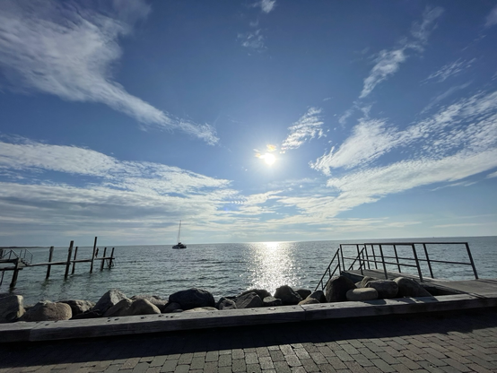 Das Bild zeigt eine ruhige Küstenlandschaft am Meer. Im Vordergrund befindet sich ein gepflasterter Weg mit einem niedrigen Holzrand, der von großen, unregelmäßig geformten Steinen gesäumt wird. Auf der rechten Seite des Weges ist ein Metallgeländer mit Stufen zu sehen, das zu einem Steg führt, der ins Meer hinausragt. Links im Bild ist ein weiterer Holzsteg mit Stufen zu erkennen. Im Hintergrund erstreckt sich das Meer, auf dem ein Segelboot ruhig schaukelt. Der Himmel ist blau mit vereinzelten weißen Wolken, und die Sonne scheint hell, wodurch ein glänzender Streifen auf dem Wasser sichtbar ist. Die Szene wirkt friedlich und idyllisch, mit einer klaren Trennung zwischen dem Land und dem Meer.