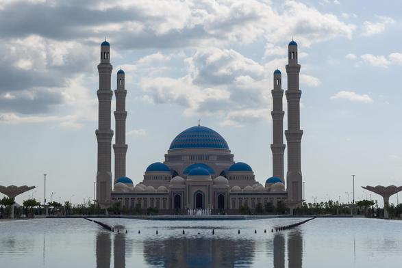The image depicts the Central Mosque of Astana, the largest mosque in Central Asia, the second largest mosque outside Middle East, and one of the largest in the world. The mosque is a large, multi-tiered structure with four towering minarets and a dome at the center. The mosque's blue color contrasts beautifully against the cloudy sky.

The mosque is situated within a large pool, adding to its grandeur. The water reflects the mosque's architectural details, enhancing the overall visual appeal of the scene. In the background, there are trees and lampposts that provide depth and context to the image.