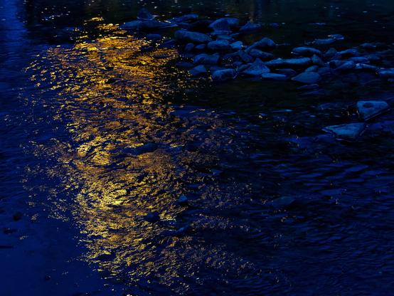 Dark blue water flows around scattered rocks with golden light reflections rippling across the surface. The twilight scene captures warm amber highlights dancing on the current while cool deep blue tones dominate the shadows between weathered stones along the riverbank.