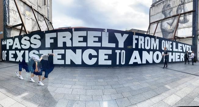A pano photo of a mural with words about being able to pass freely from one existence to another was taking with an iPhone. the people walking by are truncated due to the pano process.