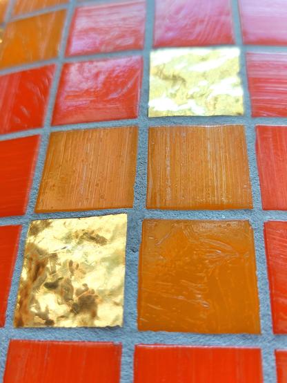 Section of a mosaic of little square glass tiles in opaque, bright orange-red and ochre-red. Some of the tiles are gilded. Grey cement can be seen in between. The tesserae have different structures: the coloured ones are flat with fine horizontal or vertical lines from the drawing of the glass. The gold-coloured ones are three-dimensional and raised. They look like tiny topographical maps or small mountains.
  // 
Ausschnitt eines Mosaiks aus quadratischen Glasplättchen in opakem, leuchtendem Orangerot und Ockerrot. Einige Steinchen sind vergoldet. Dazwischen ist grauer Zement zu sehen. Die Mosaiksteinchen haben unterschiedliche Strukturen: die Bunten sind flach mit feinen horizontalen oder vertikalen Linien vom Ziehen des Glases. Die Goldenen sind plastisch und erhaben. Sie wirken wie topografische Karten oder kleine Gebirge.