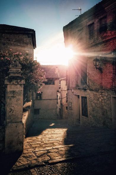 A narrow, sunlit street in a historic Spanish town. The sun flares brightly between two stone buildings, casting long shadows on the cobblestone path. A flowering oleander bush sits to the left, adding a touch of colour. The warm light and old buildings create a sense of timelessness and peace.