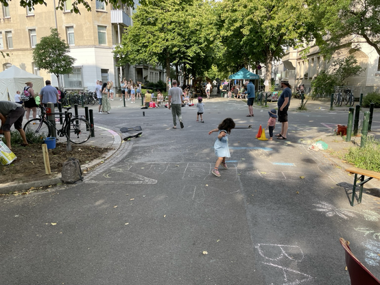 Eine Kreuzung einer Wohnstraße. Es befinden sich mehrere Menschen auf der Straße. Dabei fallen vor allem spielende Kinder auf. Weiter entfernt ist eine Tanzgruppe zu erkennen.