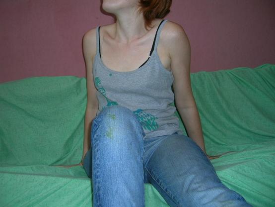 Jeune femme assise sur un canapé mou vert, devant un mur rose. Son visage est coupé. Elle porte un jean serré taché d'herbe au genou et un débardeur gris avec un pochoir fait main vert.
Le visage est tourné vers la gauche et elle semble se lever.
