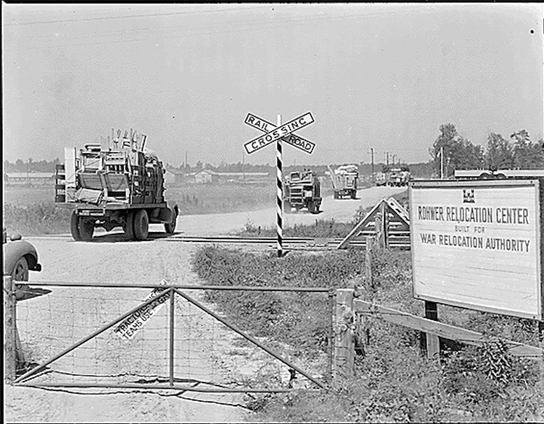 A black-and-white historical photograph showing a rural scene with a gated entrance to the Rohwer Relocation Center, operated by the War Relocation Authority. A truck loaded with furniture and household goods is passing through the gate, with additional trucks visible in the background. A railroad crossing sign with "RAILROAD CROSSING" is positioned near the gate, and a sign reading "ROHWER RELOCATION CENTER WAR RELOCATION AUTHORITY" is prominently displayed on the right side. The landscape includes open fields and distant trees under a clear sky.