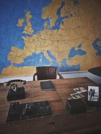 A high-angle photograph shows a large wooden desk with a brown leather chair behind it. On the desk are various items, including an old-fashioned black rotary telephone on the left, a notepad in the center, and a set of old black and white photographs on the right. A large map of Europe is visible on the wall in the background. The photo is well-lit and has a vintage aesthetic.