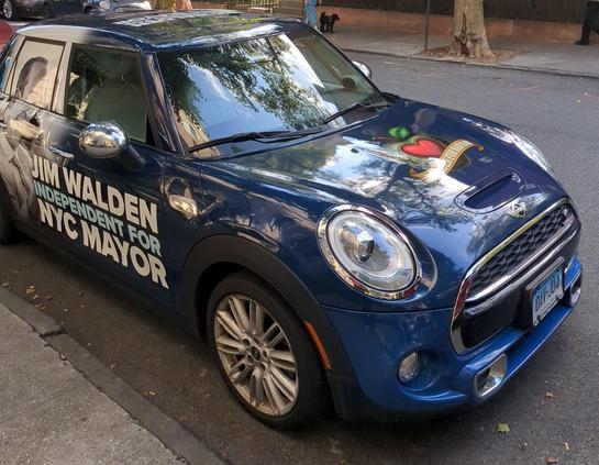 Photograph of Jim Walden's NYC Mayor campaign car. It is a mini cooper parked by the sidewalk in Brooklyn Heights. It is painted dark blue with a grayscale photograph of a smiling Jim Walden painted on the rear door. On the side of the front door we see JIM WALDEN INDEPENDENT FOR NYC MAYOR. On the hood is a campaign logo of a heart-shaped apple with a "WALDEN FOR MAYOR BANNER." The car has a Connecticut license plate.