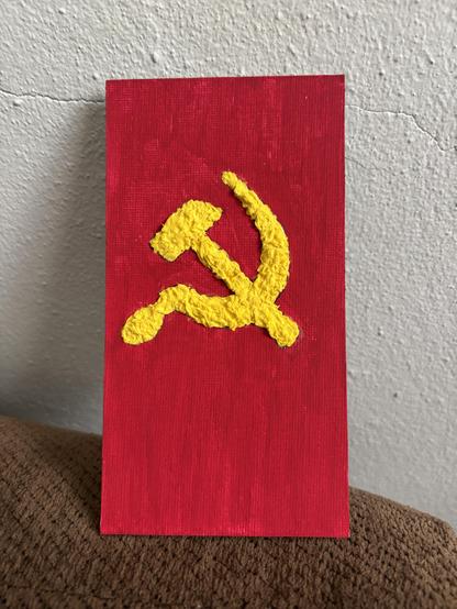 Martillo y Hoz realizado con papel maché en un canvas de tamaño 3x6 pulgadas. El canvas está pintando de color rojo y la silueta, que es al relieve, del martillo y la hoz está realizado en color amarillo.