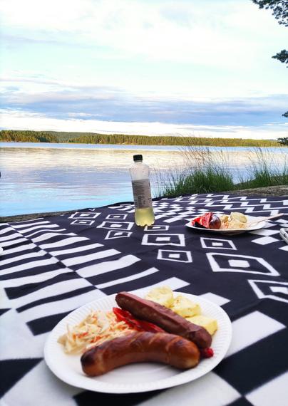 Kaksi lautasellista ruokaa ja juomapullo viltillä kalliolla. Taustalla järvi ja järven takana metsänraja.