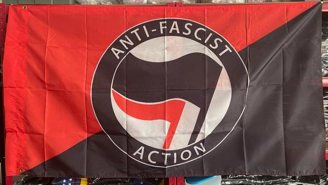 Eine Flagge der Antifaschistischen Aktion mit rot-schwarzem Hintergrund, einem Kreis mit "ANTI-FASCIST ACTION" beschriftet und im Kreis das Logo der Antifa mit der roten hinter der schwarzen Flagge stilisiert.