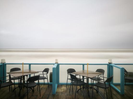 das bild zeigt eine langzeitbelichtung des meeres von der außenterrasse eines strandrestaurants aufgenommen.
die horizontlinie trennt das bild in eine obere und eine untere hälfte. 
oben ist der graue, bedeckte wolkenhimmel zu sehen. darunter ist das meer zu sehen. es ist sehr aufgewühlt und auch stürmisch, doch durch die langzeitbelichtung ist auch das meer weichgezeichnet.
unten ist die terrasse zu sehen. tische mit jeweils vier schwarzen stühlen stehen dort. die terrasse hat eine hellblaue balustrade und einen holzboden. es ist nassgeregnet.
die szene wirkt ruhig und leer, es ist niemand auf den sitzen zu sehen. das gesamte bild ist in eher kalten, gedämpften tönen gehalten.