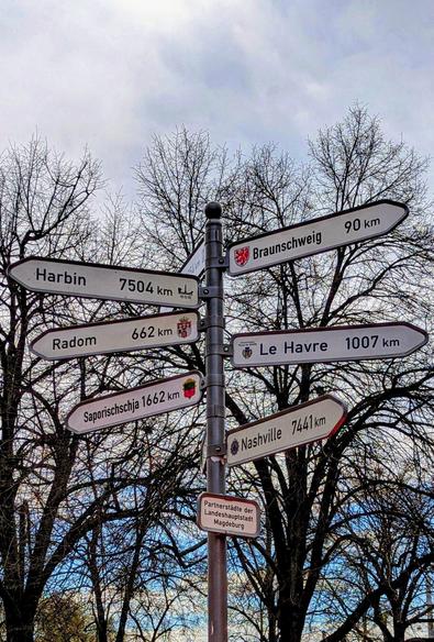 A metal signpost in Magdeburg, Germany, with white directional signs pointing to its twin towns. From top to bottom on the left, the signs read: Harbin 7504 km, Radom 662 km, and Saporischschja 1662 km. On the right, they point to Braunschweig 90 km, Le Havre 1007 km, and Nashville 7441 km. A smaller sign at the base says 'Partnerstädte der Landeshauptstadt Magdeburg' (Twin towns of the state capital Magdeburg). The signpost is in front of bare trees and a cloudy sky.