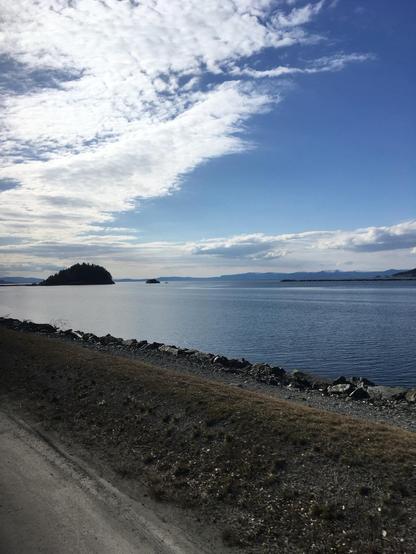 A serene body of water reflects a partly cloudy sky, with a small island and distant hills visible in the background. The foreground features a rocky shoreline and a grassy bank.