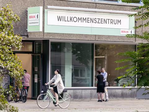 Aufnahme des physischen Willkommenszentrums in Berlin, zwei Menschen stehen davor und unterhalten sich. Eine Radfahrerin fährt vorbei.