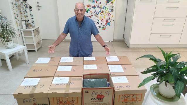 גבר מבוגר בחולצה כחולה עומד מאחורי שולחן עם 12 קרטונים חומים מסודרים בשורות. הקרטונים ממוספרים מ-1 עד 12 ועליהם מודבקים דפים לבנים. הגבר מחייך ופורש את ידיו בתנועה מזמינה. ברקע נראים ארונות מטבח לבנים, צמחים ירוקים, ועל המקרר מגנטים צבעוניים. הסביבה נראית כמו מטבח או אזור מגורים ביתי.