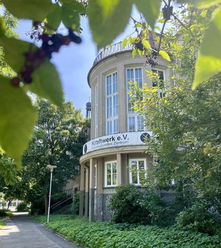 Blick auf ein historisches Gebäude mit einem aus grünem Gebüsch herausragenden Rondell mit der Aufschrift Kraftwerk e.V. Soziokulturelles Zentrum