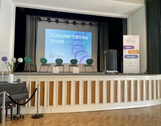 Blick auf eine Bühne mit fünf grünen Stühlen, dahinter sieht man groß ZUKUNFTSTAG 2025