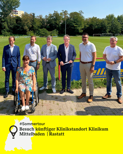 Sieben Personen, darunter Minister Manne Lucha und Landes-Behindertenbeauftragte Nora Welsch, stehen vor einem Fußballfeld mit Toren und Bäumen im Hintergrund. Unten gelber Balken mit Text: Sommertour. Besuch künftiger Klinikstandort Klinikum Mittelbaden  | Rastatt.