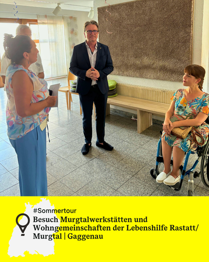 Minister Manne Lucha spricht mit drei Personen in einem hellen Raum mit Sitzbank und Fenster. Rechts neben ihm im Rollstuhl ist die Landes-Behindertenbeauftragte Nora Welsch. Links steht eine Frau. Text: Sommertour. Besuch Murgtalwerkstätten und Wohngemeinschaften der Lebenshilfe Rastatt/ Murgtal | Gaggenau.