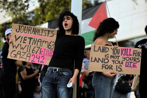 Two people holding up cardboard signs, wearing jeans, dark tops, and have dark brown long curly hair. The person on the left is shouting out towards the street. Their sign says "Journalists / Vietnam 63 / WWII 67 killed / Gaza 242". The one on the right is facing down with their sign stating "Land you kill for is not yours / Land you die for is".

The background is filled with people at the rest of the protest. A Palestine flag is framed in the center behind the subjects.