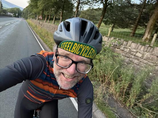 A smiling cyclist wearing glasses and a helmet poses for a selfie on an empty road. The cyclist is dressed in a colorful cycling jersey and a headband with the text "STEELBICYCLE." The background features trees and a stone wall.