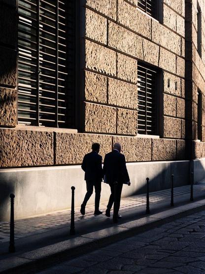 Two men in business suits walk on a sidewalk as the low light of sunset creates long, hard shadows against the building's facade on their left. Their silhouettes create a stark contrast against the lit wall.