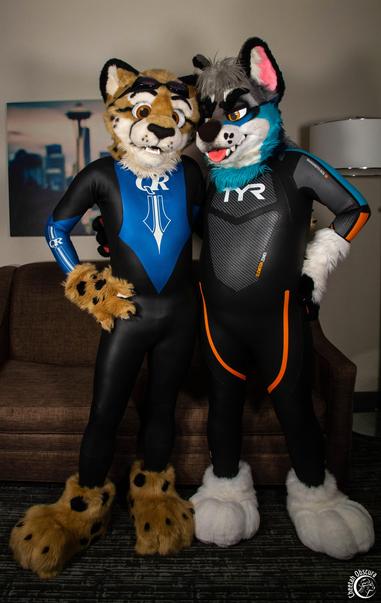 Fursuit photo of Obscura and Morro standing in a hotel room. Obscura is wearing a blue and black QR wetsuit and Morro wearing a black, orange, and blue TYR wetsuit