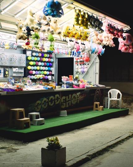 A shooting stand with lots of plush animals hanging around. A plastic chair at the front. The intense light of the stands covers the scene. The front panel says "good shot"