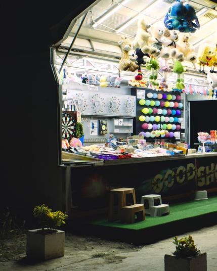A shooting stand with lots of plush animals hanging around. A plastic chair at the front. The intense light of the stands covers the scene. The front panel says "good shot"