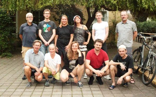 Eine Gruppe von 11 Menschen steht und hockt vor der Kamera. Eine duerchaus diverse Gruppe. Leider nicht vollständig.