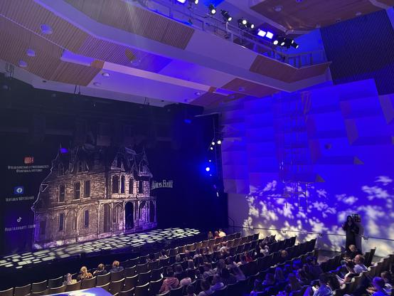The image shows a theater stage set for a performance of "The Addams Family." There is a detailed backdrop of a gothic house, dim lighting, and an audience seated in rows. The stage has blue and purple lighting, creating a dramatic atmosphere.