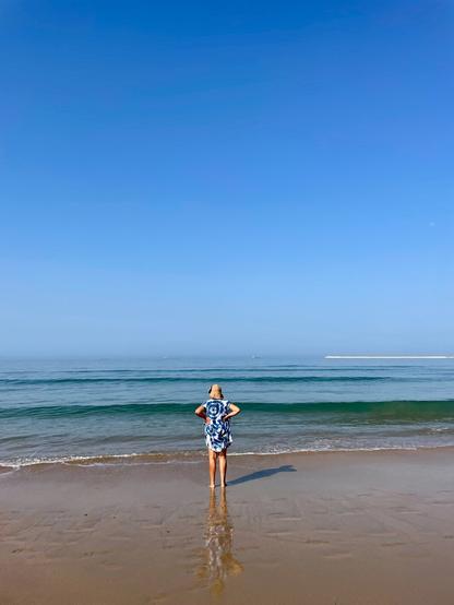 A person stands on a sandy beach, looking out at the calm sea under a clear blue sky. The individual is wearing a blue and white patterned outfit and a hat, with their hands on their hips, reflecting on the water's surface.
