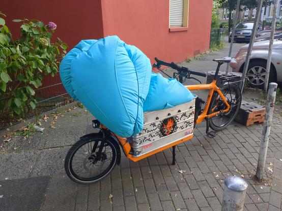 Ein Lastenrad mit großer Aluminiumkiste und zwei großen Sitzsäcken geladen. Der eine ist größtenteils in der Kiste, der andere ist vorne festgebunden so dass er über das Vorderrad hinusragt und ein gutes Stück höher ist als der Lenker.